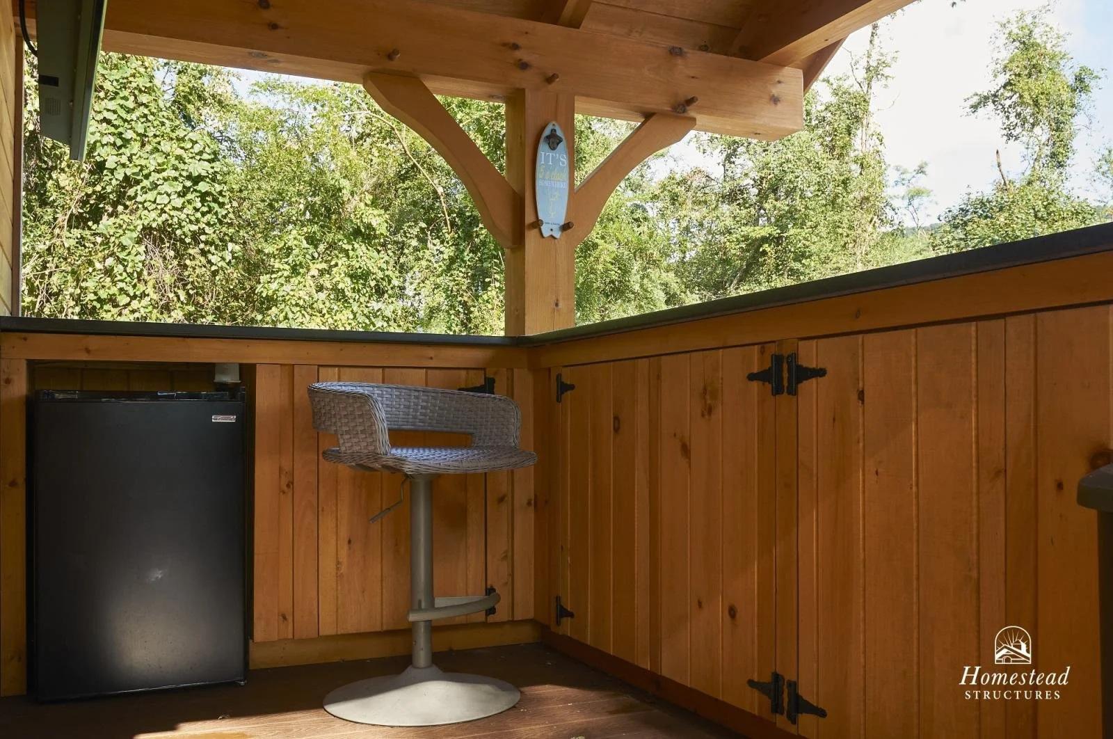 Wooden porch area with black mini fridge, wicker swivel chair, and view of trees outside, with a hanging surfboard decor on the support beam and a logo for Homestead Structures in the bottom right corner.