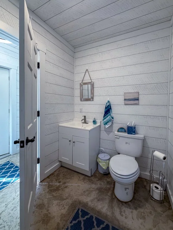 Small bathroom with white shiplap walls and ceiling, a white vanity with a sink, a framed mirror hanging above, a towel hook with a blue towel, a toilet with a box of tissues and a toilet paper holder nearby, and a small trash bin. There are two blue