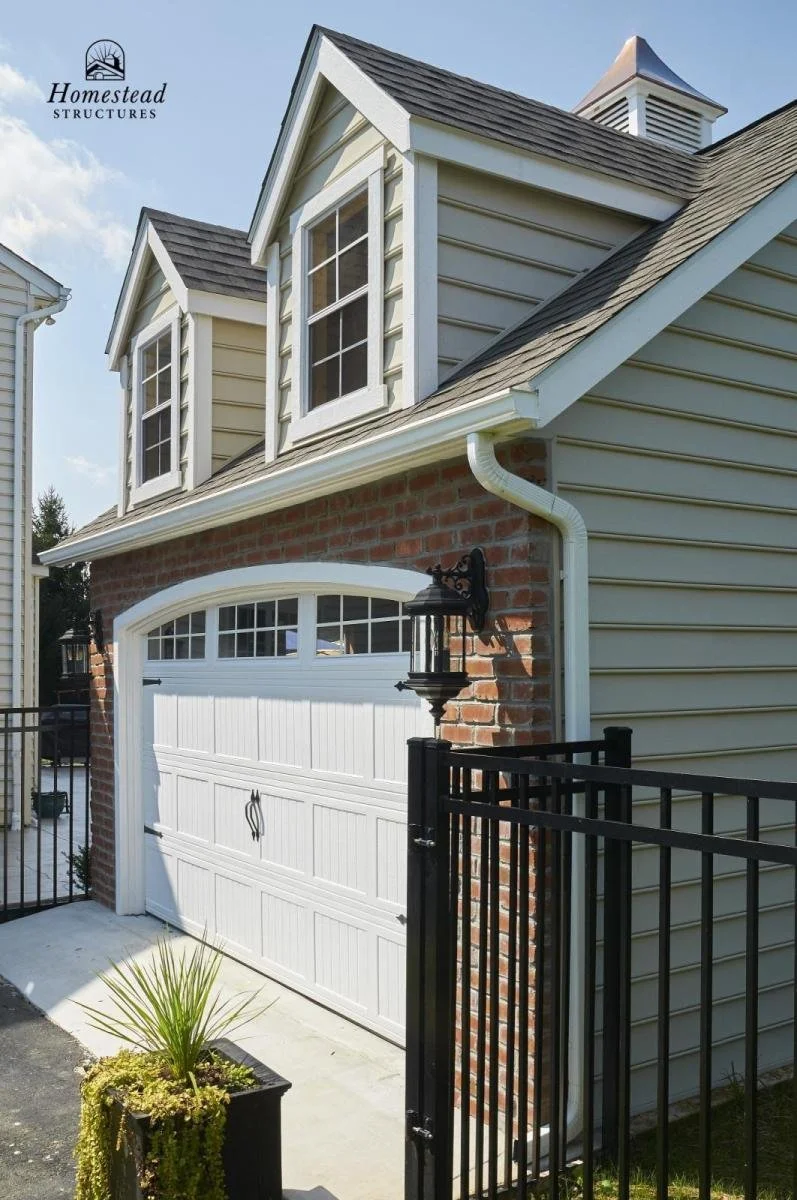 Front of a house with a white garage door, brickwork, and beige siding with three dormer windows. There is a black metal gate and a small potted plant in the foreground. A black outdoor lantern is mounted on the brick wall.