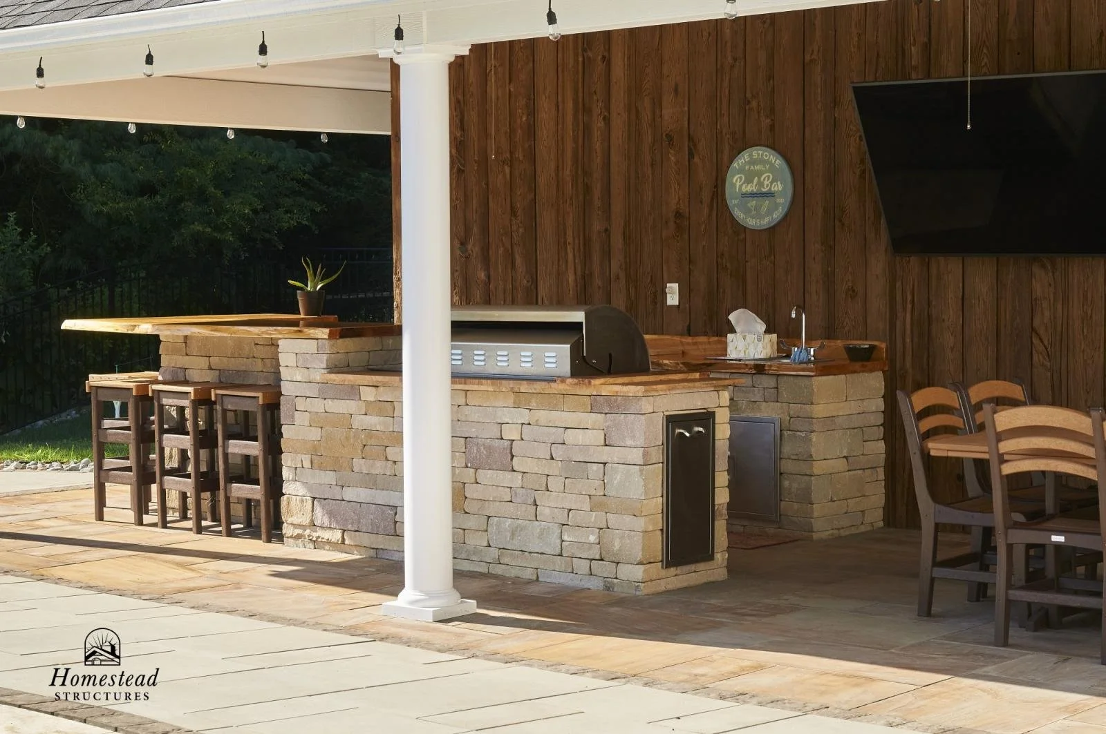 Outdoor bar area with a stone counter, bar stools, wooden chairs, a fish tank on wall, and string lights, part of a homestead structure.