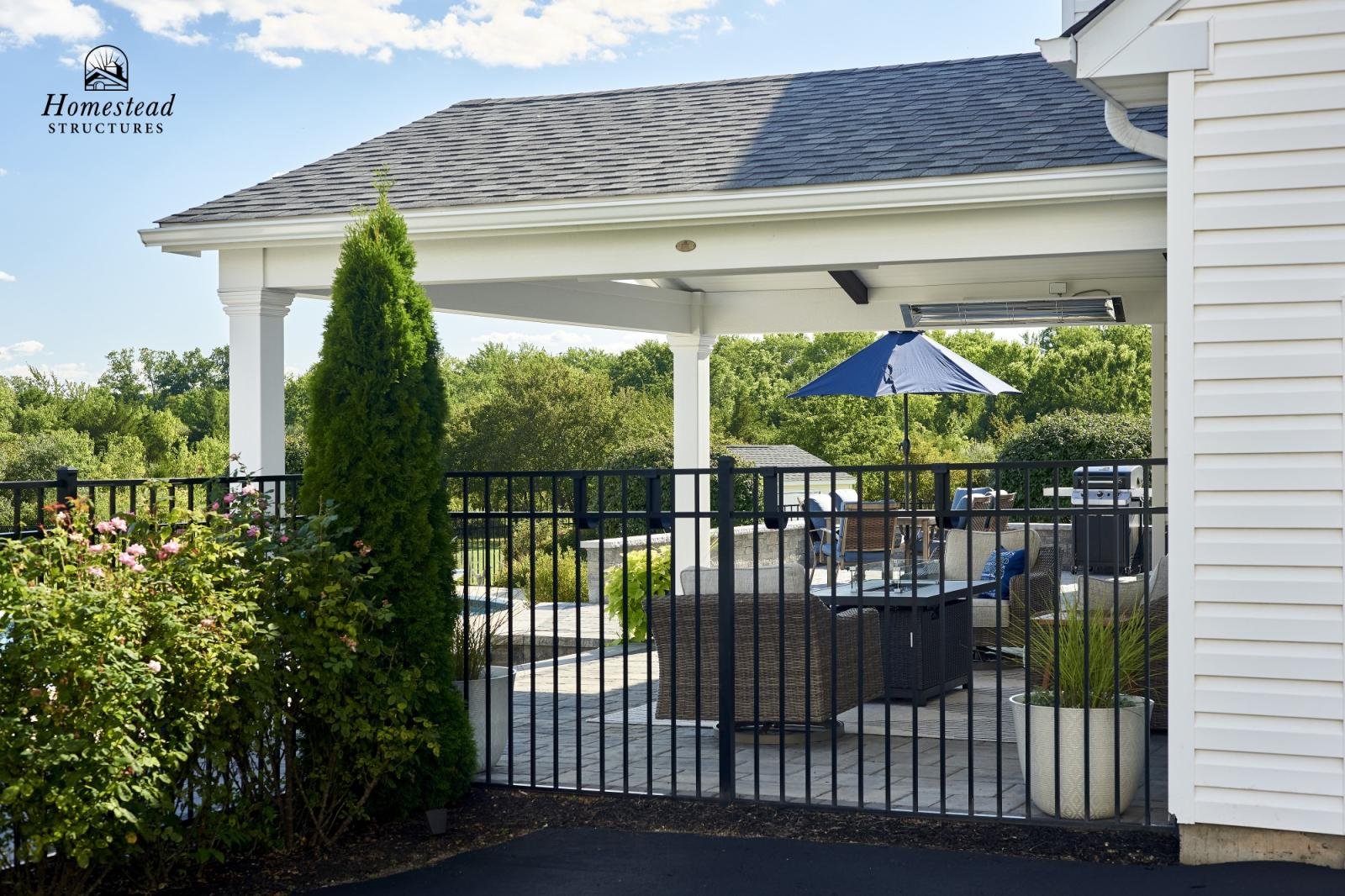 Outdoor patio area with seating, table, umbrella, and grill, enclosed by a black metal fence, attached to a white house with a porch, surrounded by green trees.