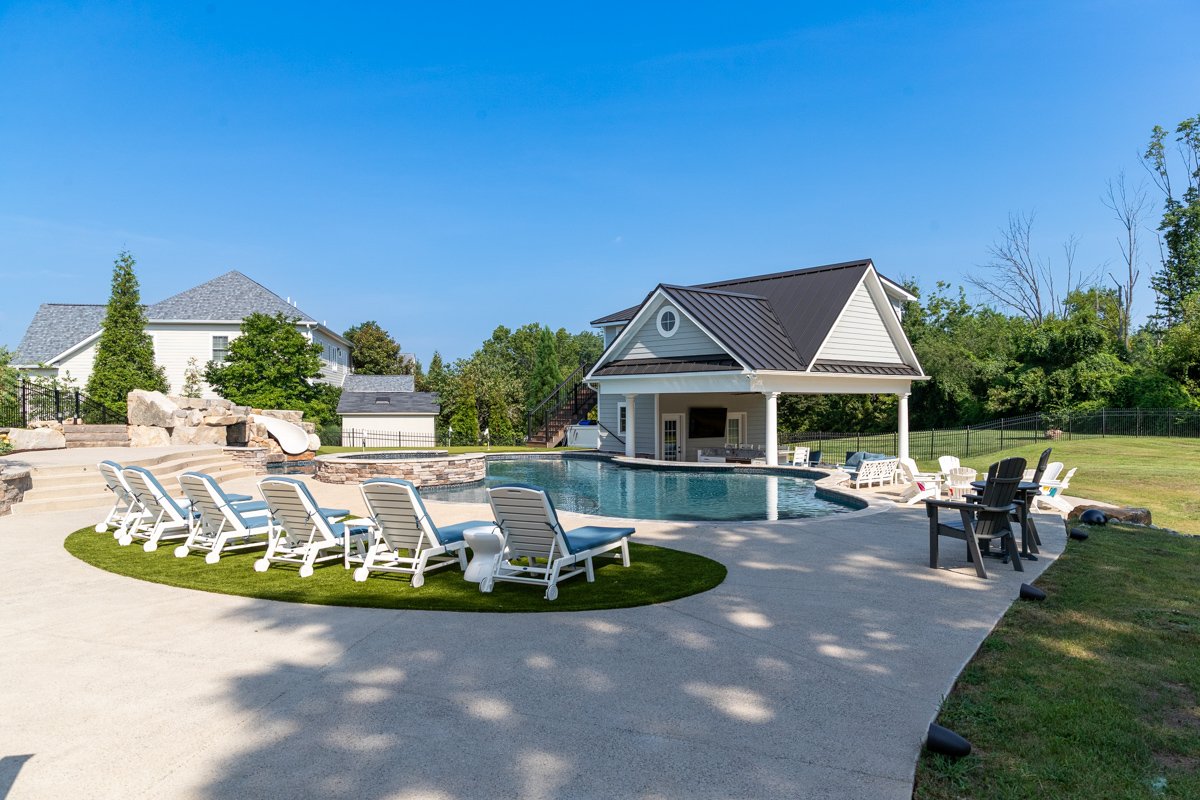 Residential backyard with a swimming pool, poolside chairs, and a pool house surrounded by trees and houses under a clear blue sky.