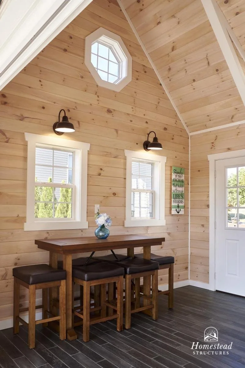 Wood-paneled wall with three black sconces, two windows, an octagonal window, a small wooden table with four stools, a vase with flowers, and a decorative wall sign with sayings about family, the anchor, and life's storms.