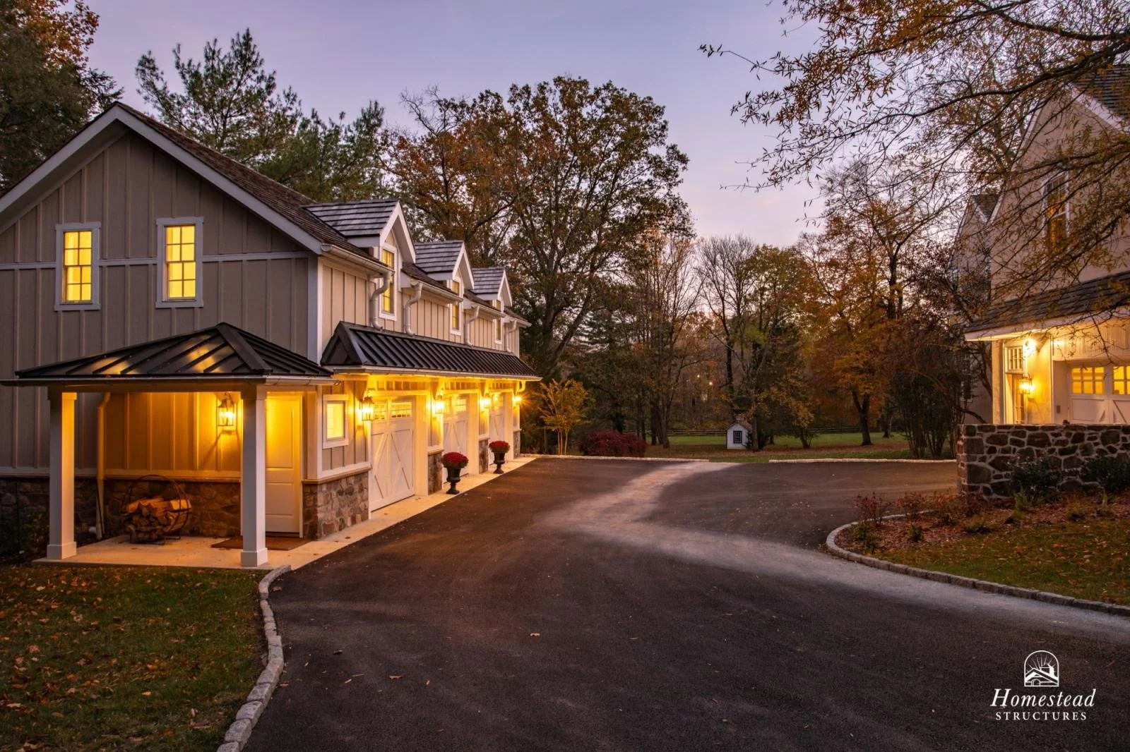 A large residential property with a driveway, multiple garage doors, exterior lighting, and a small shed in the background surrounded by trees during dusk.