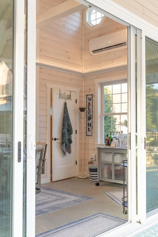 View through sliding glass doors into a bright, cozy sunroom with light wood-paneled walls, a white door with a towel hanging on it, a 'Welcome' sign hanging next to the door, a window showing trees outside, a small cabinet with bottles on top, and a