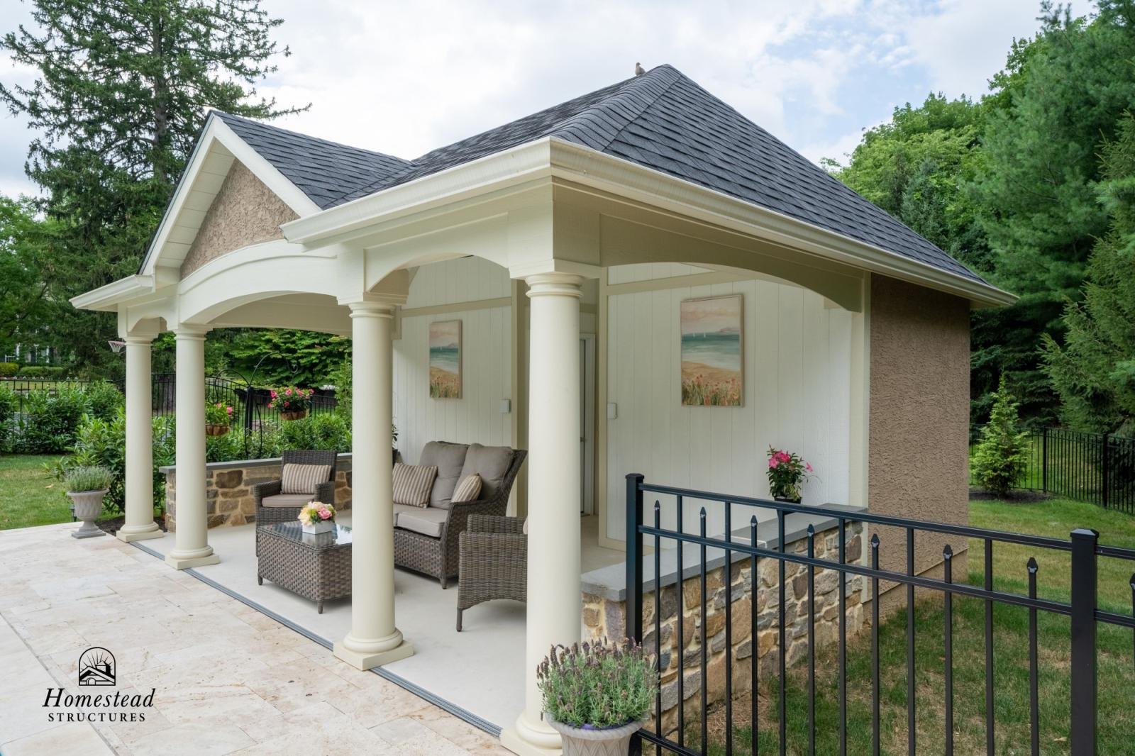 A small, elegant house with a porch supported by white columns, decorated with outdoor furniture and potted plants, surrounded by green trees and a grassy yard.
