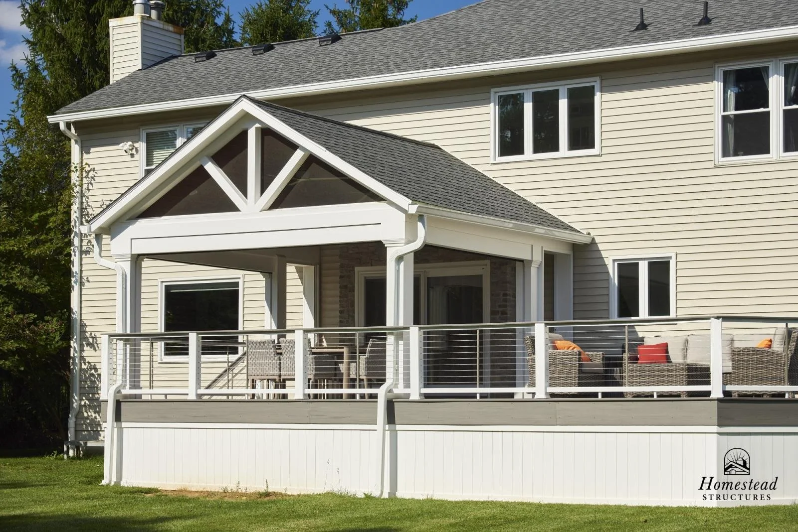 Back view of a beige two-story house with a newly built front porch with white railing, wicker chairs, and outdoor furniture, surrounded by a green lawn.