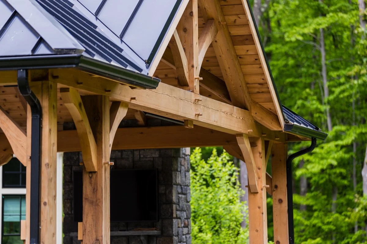 Close-up of a wooden house under construction with a metal roof and green trees in the background.
