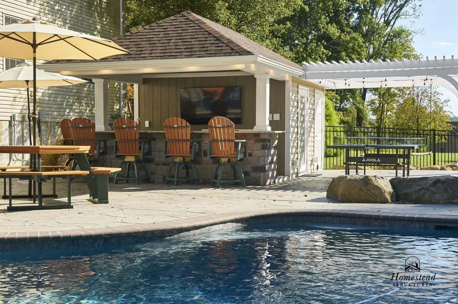 Backyard pool area with outdoor bar, seating, umbrellas, and ping pong table, surrounded by trees and fencing.