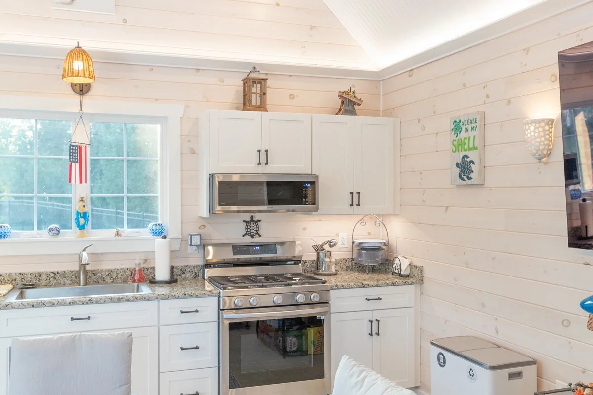 Kitchen with white cabinets, granite countertops, stainless steel stove and microwave, window with American flag and nautical decor, wall art saying 'Eat EASE IN MY SHELL', and some decorative items.