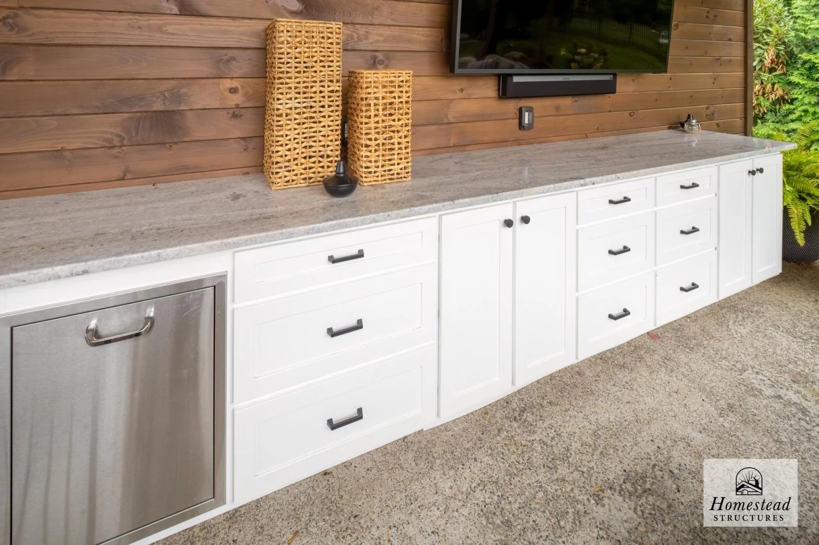 Outdoor kitchen with white cabinetry, black handles, a stainless steel mini-fridge, a granite countertop, and a wooden wall background. Decor includes wicker boxes and a vase, with a TV mounted on the wall and a potted fern plant nearby.