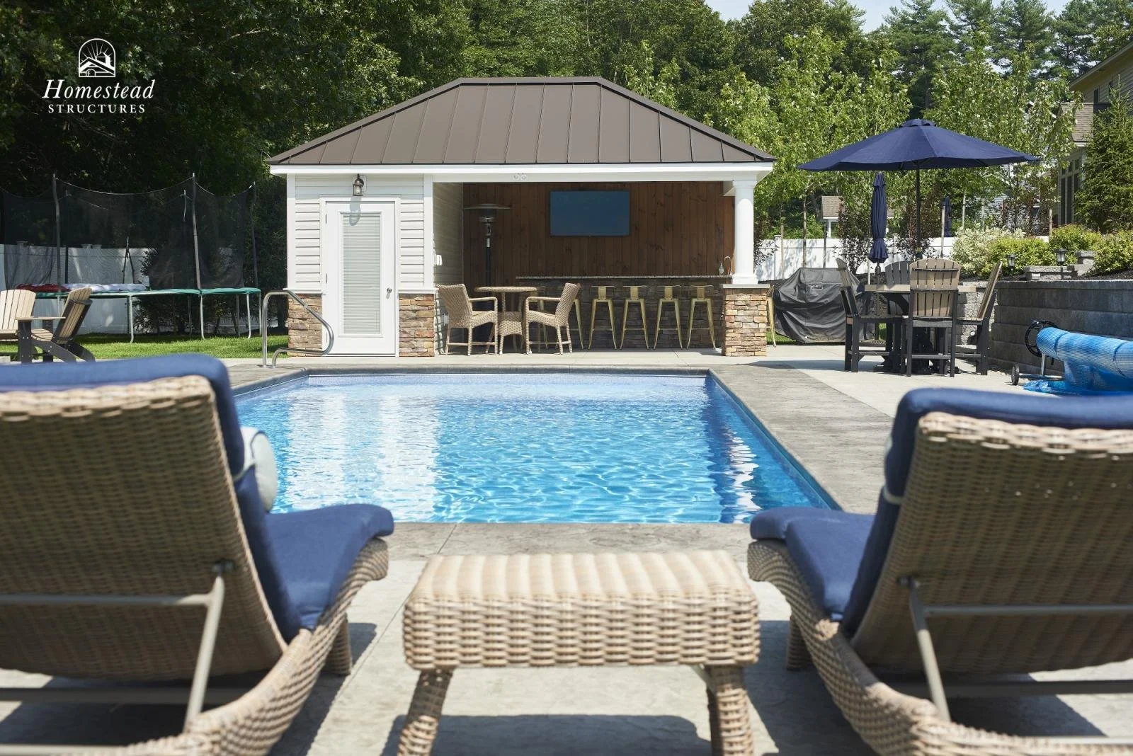 Backyard with swimming pool, poolside chairs, and a pool house with outdoor kitchen and dining area under a shade umbrella.