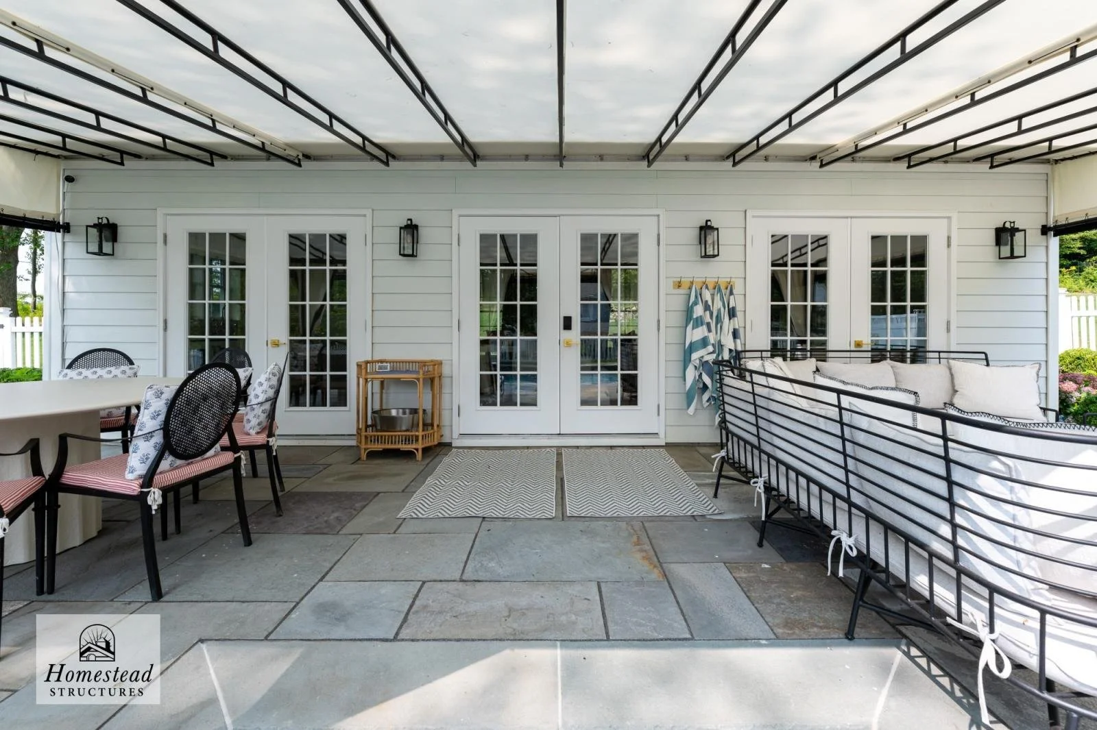 Covered outdoor patio area with a white house exterior, French doors, black wall-mounted lanterns, outdoor furniture including chairs and a dining table, striped towels hanging on a wall rack, and a decorative black metal bed frame with pillows.
