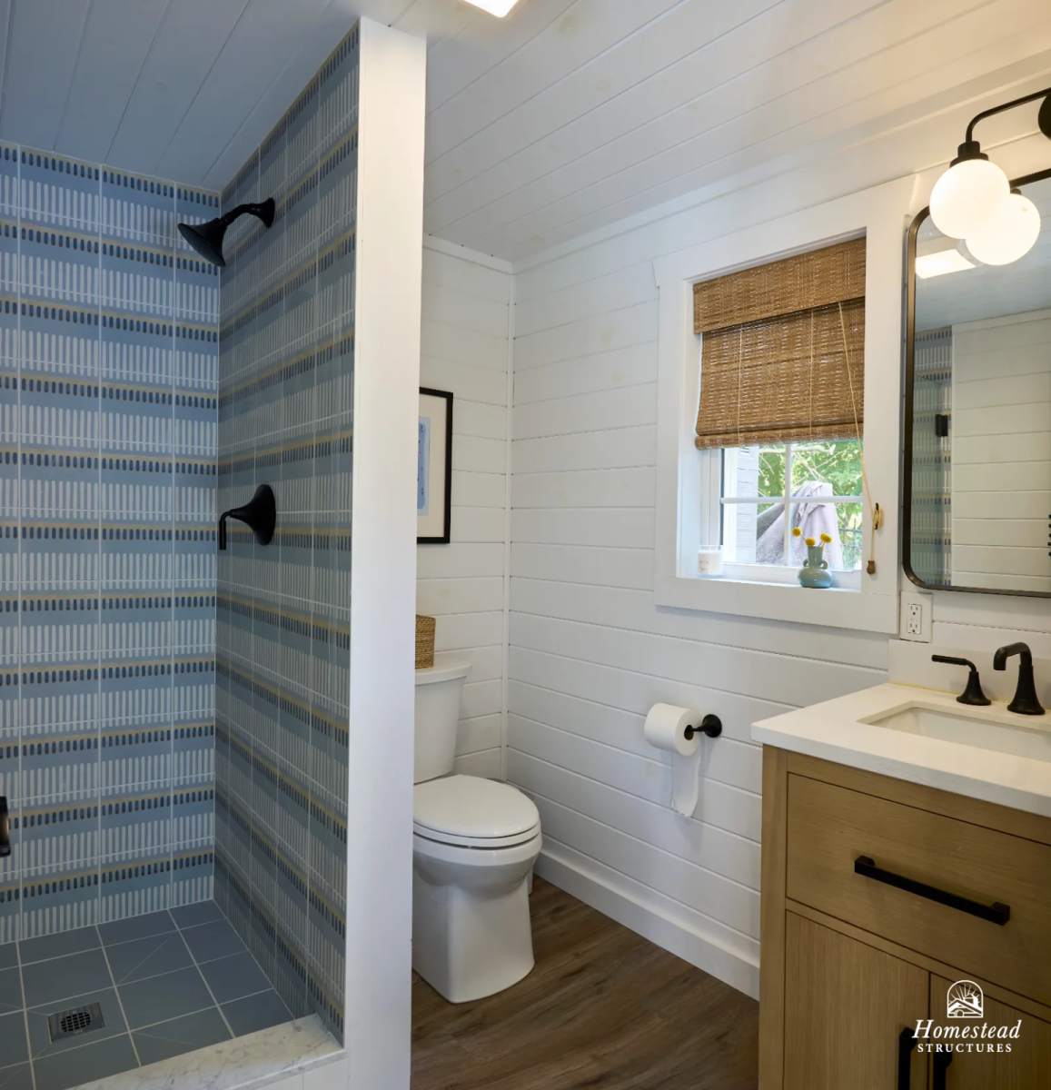 Bathroom with a walk-in shower featuring blue patterned tile and black fixtures, a toilet with a basket on top, a window with a woven shade, a small vanity with a light wood cabinet, a white counter, and a mirror with black hardware.