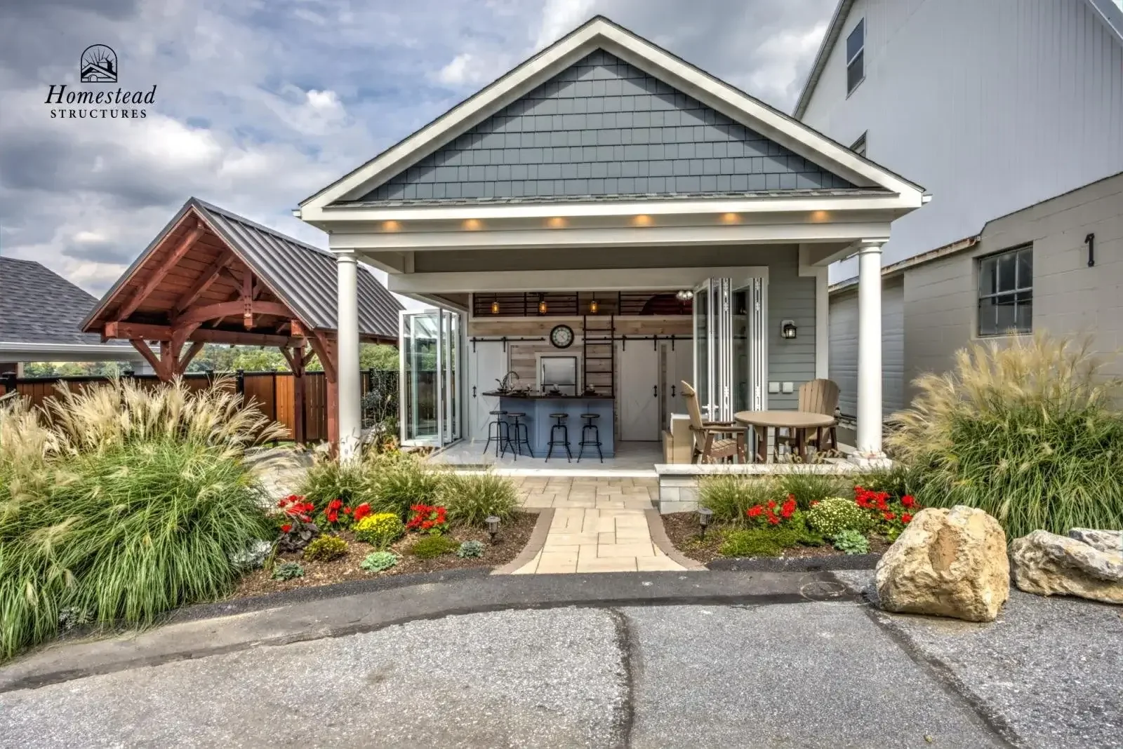 View of a backyard patio with a covered outdoor kitchen and seating area, landscaping with bushes and flowers, and large rocks in the foreground.