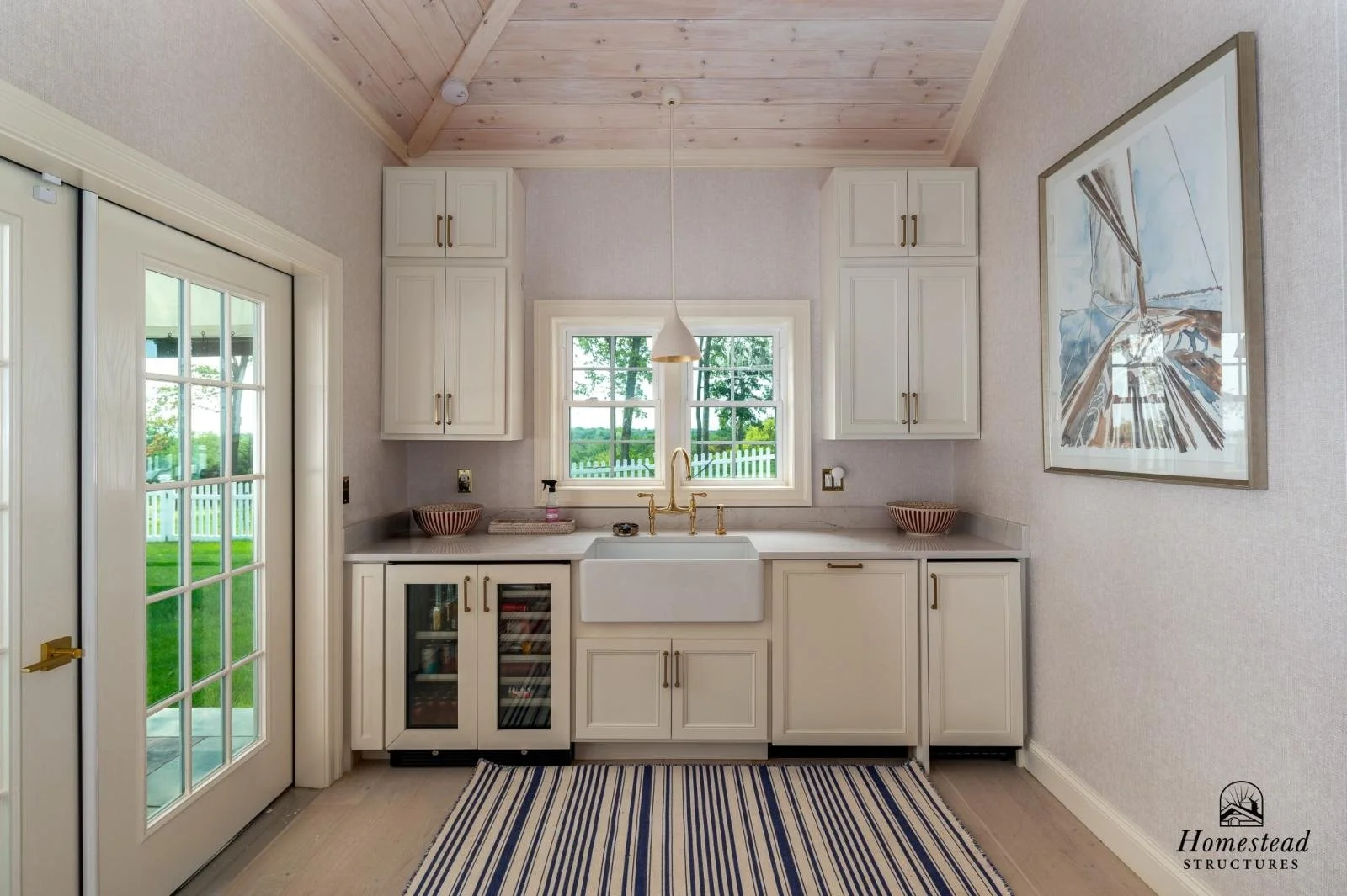 A bright kitchen with white cabinetry, a farmhouse sink, a small window above the sink, and French doors leading outside. There is a striped rug on the floor and a large painting on the right wall. The ceiling is wood-paneled.