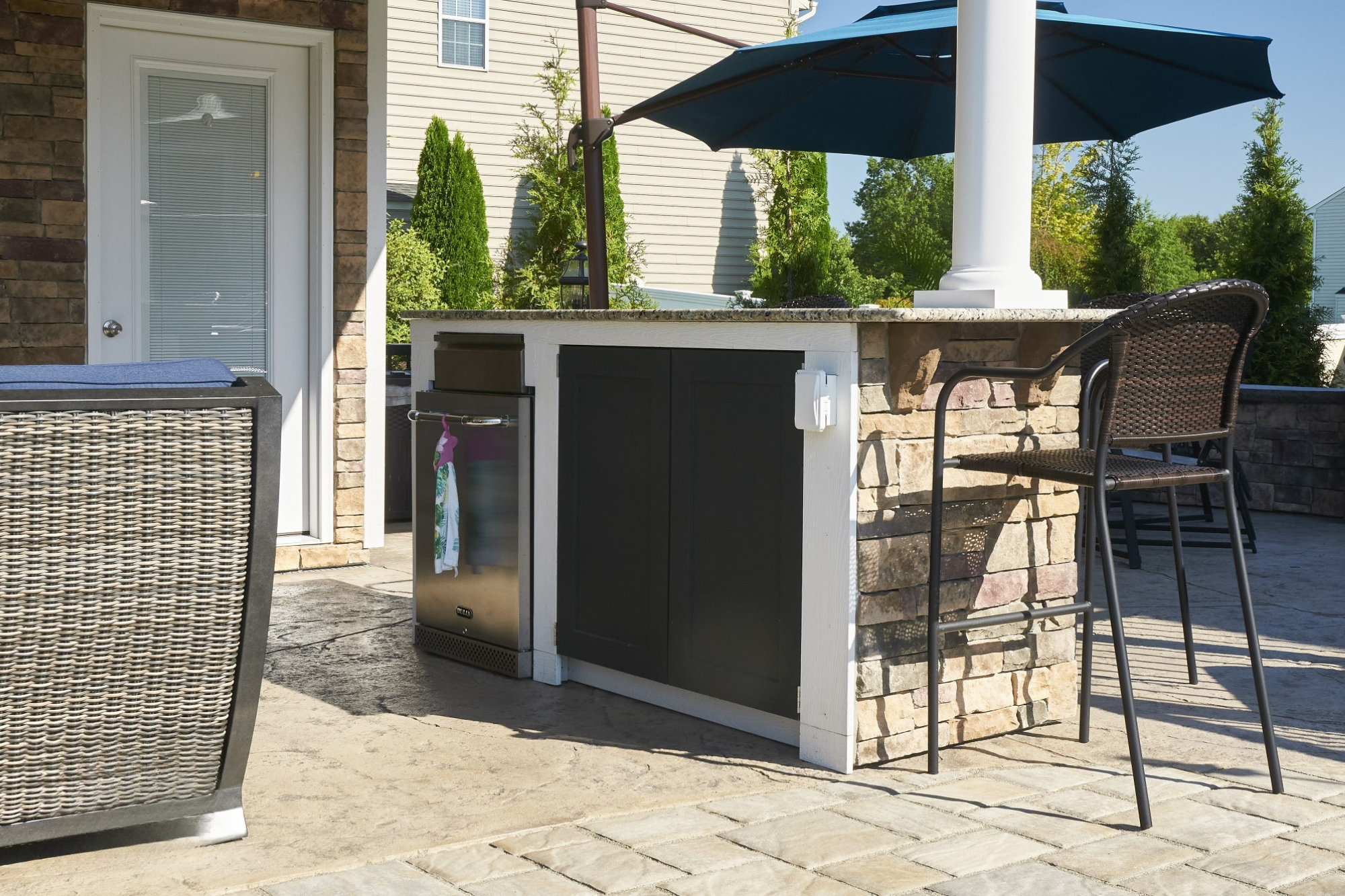 Outdoor kitchen area with a small refrigerator, a counter with a large umbrella, wicker chairs, located on a stone patio with greenery in the background.