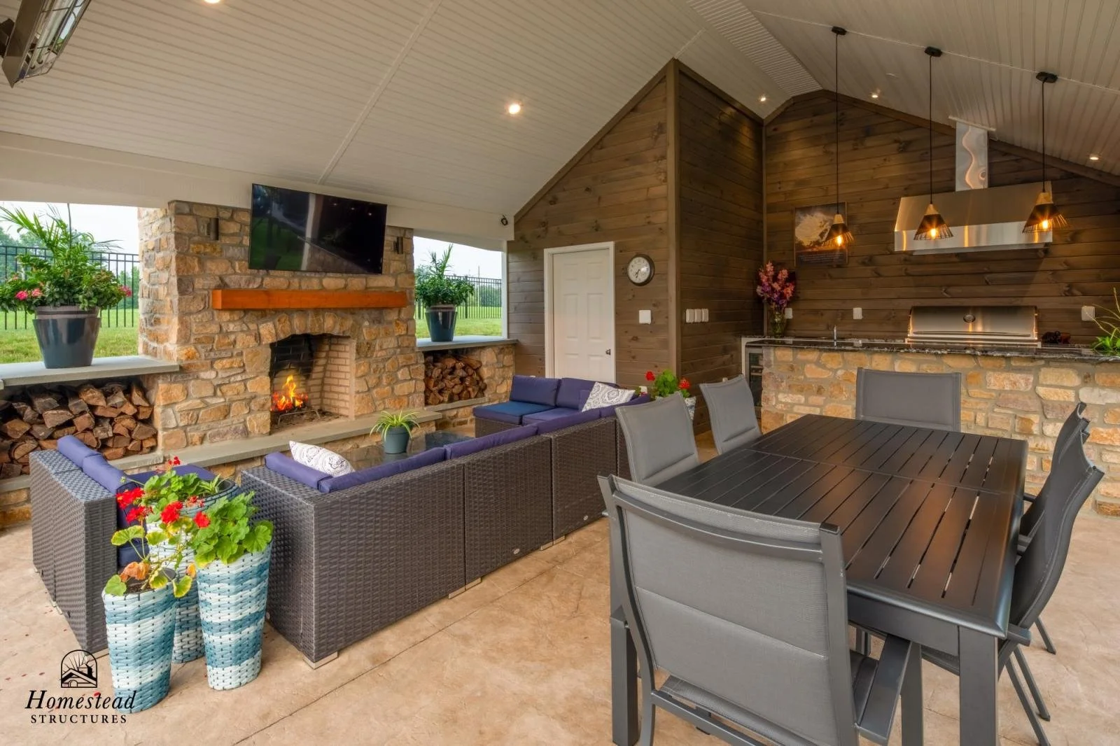 Covered outdoor patio with a brick fireplace, wicker sofas with purple cushions, a black dining table with six gray chairs, potted flowers, wood accents, and a mounted TV.