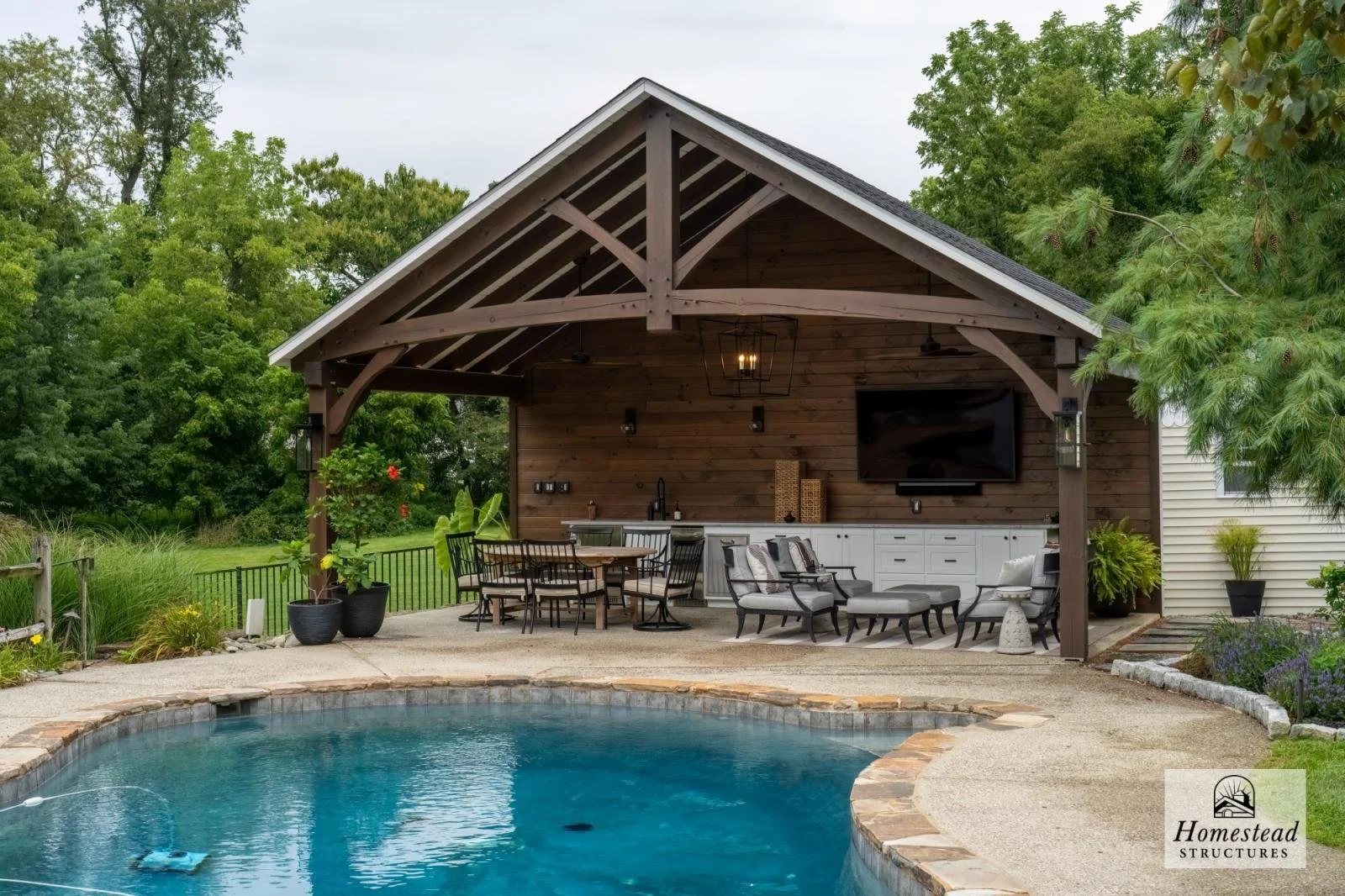 Backyard with swimming pool, outdoor seating area, and a wooden pavilion with a mounted TV, surrounded by greenery.