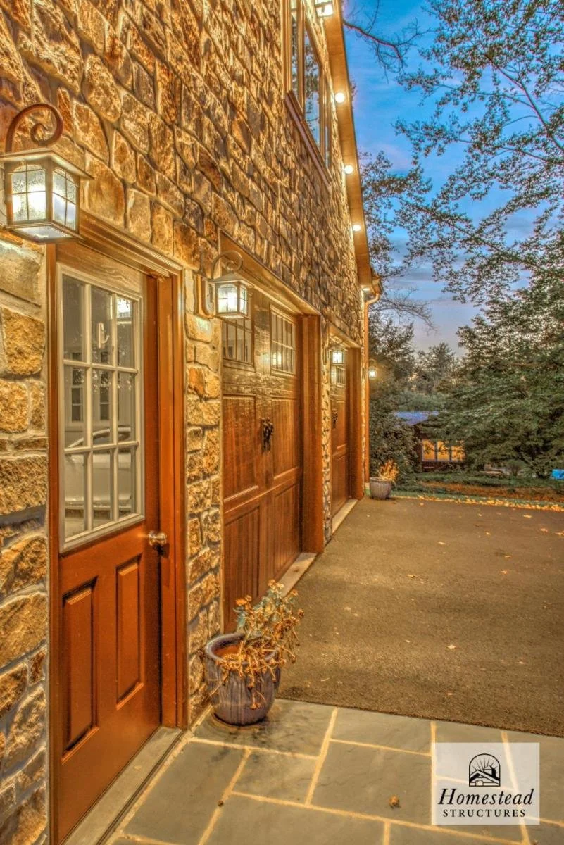 Exterior view of a stone house with wooden doors, metal lanterns near the door, and warm interior lighting visible through the windows. A potted plant sits on the stone porch, and trees with leaves are visible in the background.