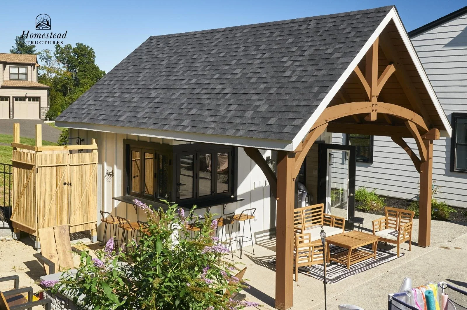 Backyard of a small building with a gray shingle roof, black window frames, and a wooden covered porch with outdoor seating, surrounded by plants and a gravel patio.