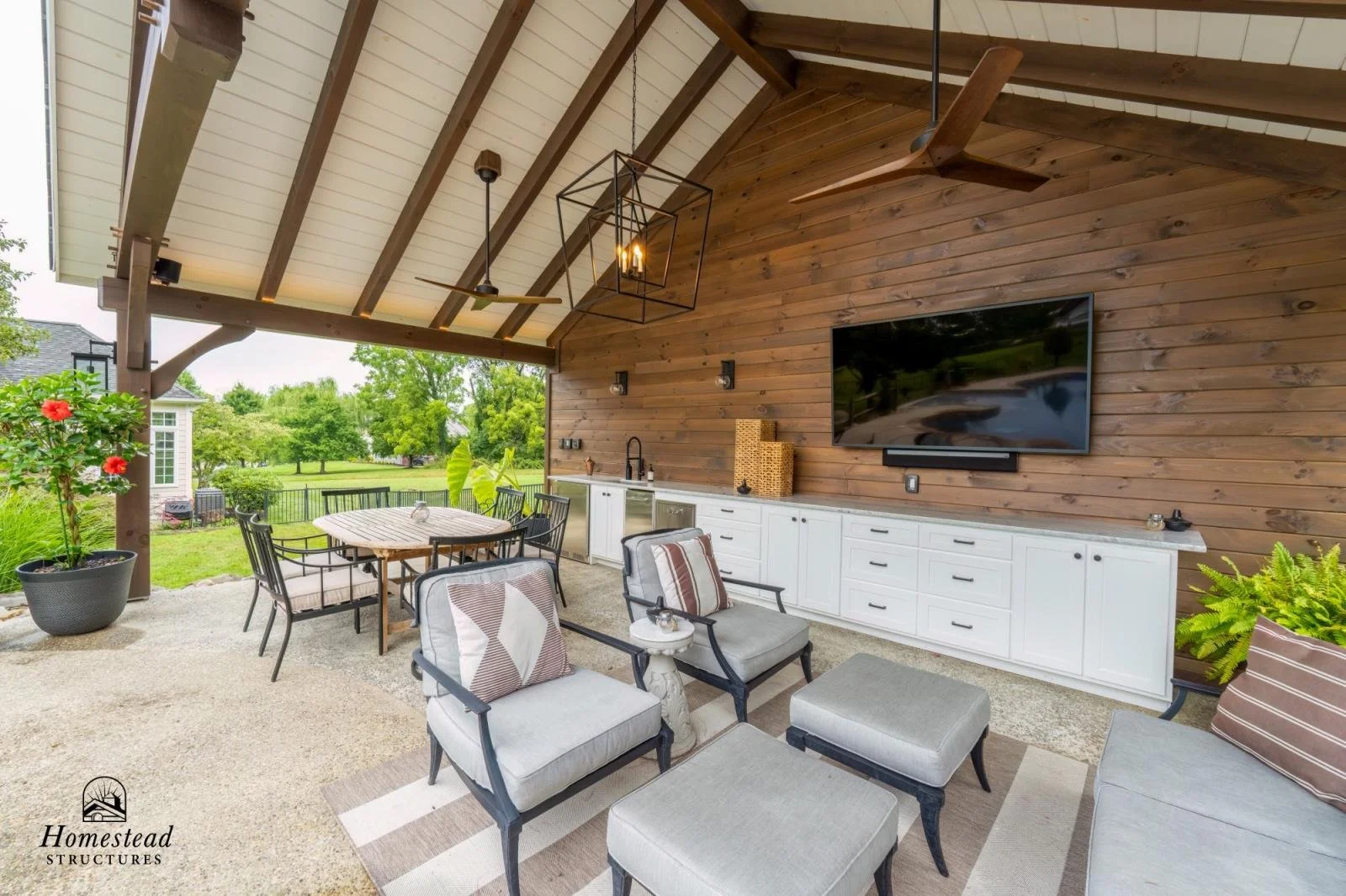 Outdoor living space with a wood-paneled ceiling, ceiling fan, mounted flat-screen TV, white storage cabinet, seating area with upholstered chairs and ottomans, a dining table with chairs, and potted plants, overlooking a grassy yard with trees.