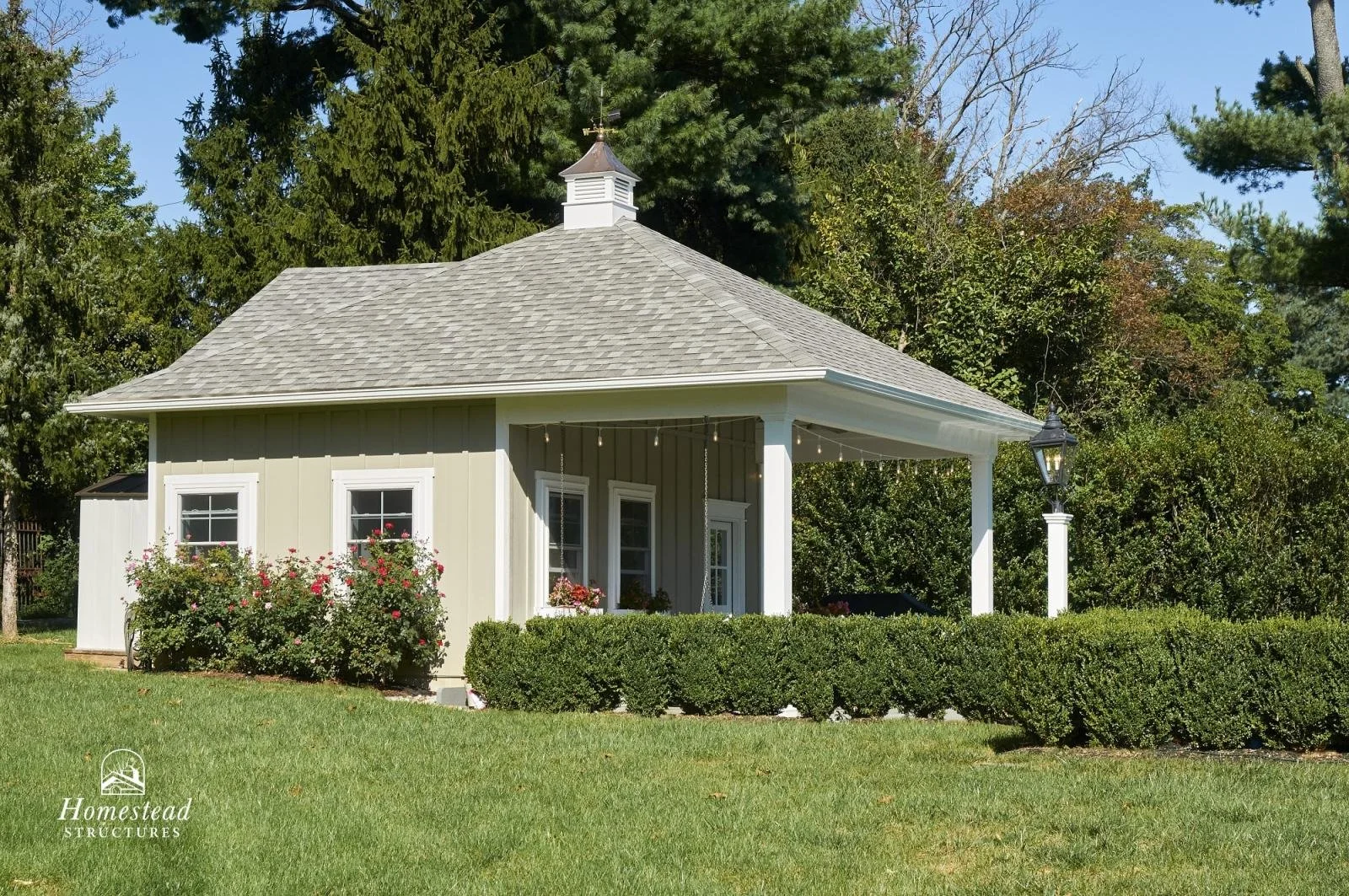 Small, light green cottage with white trim, three small windows with flower boxes, a decorative cupola on the roof, and a black lantern style lamp on the porch.