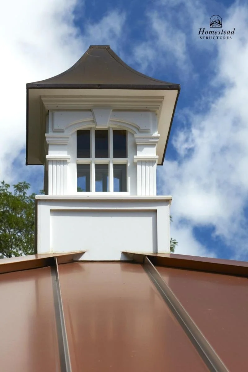 A decorative white architectural detail with a window and columns on top of a building, viewed from below against a partly cloudy sky.