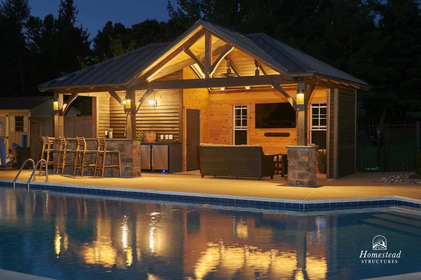 A wooden poolside cabana with outdoor seating, bar, and a swimming pool reflecting lights, at dusk.