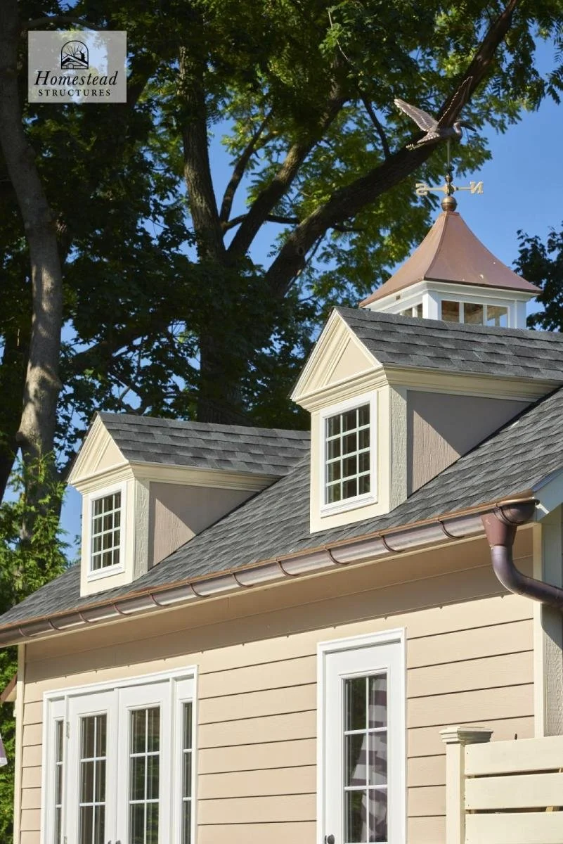 Close-up of a beige house with two dormer windows, gray shingle roof, white trim, and a weather vane with a bird on top, under a large tree with green leaves and a blue sky.
