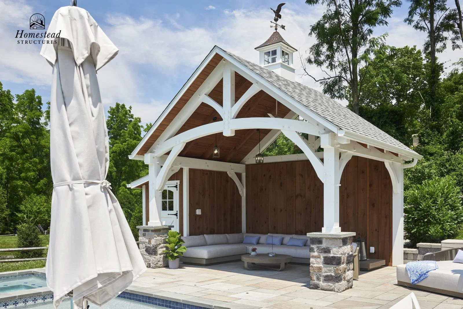 Outdoor poolside patio with a white canopy structure, a sectional sofa, potted plants, and a closed umbrella, surrounded by green trees under a blue sky.
