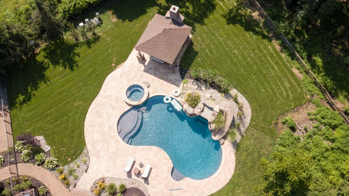 Aerial view of a backyard with a swimming pool, hot tub, and a small pavilion surrounded by grass and trees.