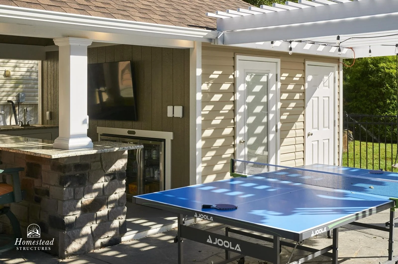 Backyard patio area with a ping pong table, a mounted flat-screen TV, and a small outdoor kitchen with a counter and sink. The area is partially shaded by a pergola with string lights. There are two white doors and green trees in the background.