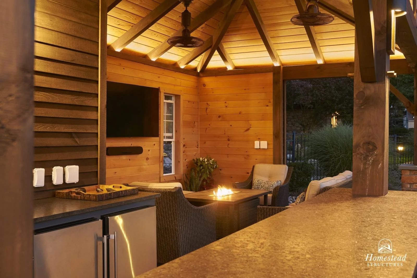 Outdoor patio with wooden walls and ceiling, a gas fireplace, wicker chairs, and a small refrigerator, illuminated by ambient lighting.