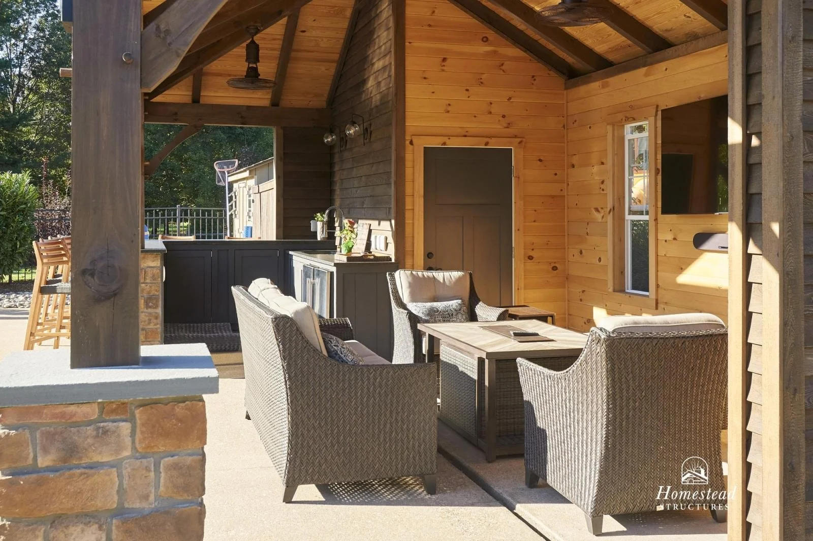 Outdoor covered patio area with wicker chairs and a wooden table, adjoining a kitchen with black cabinetry, a sink, and wood paneling on the walls.