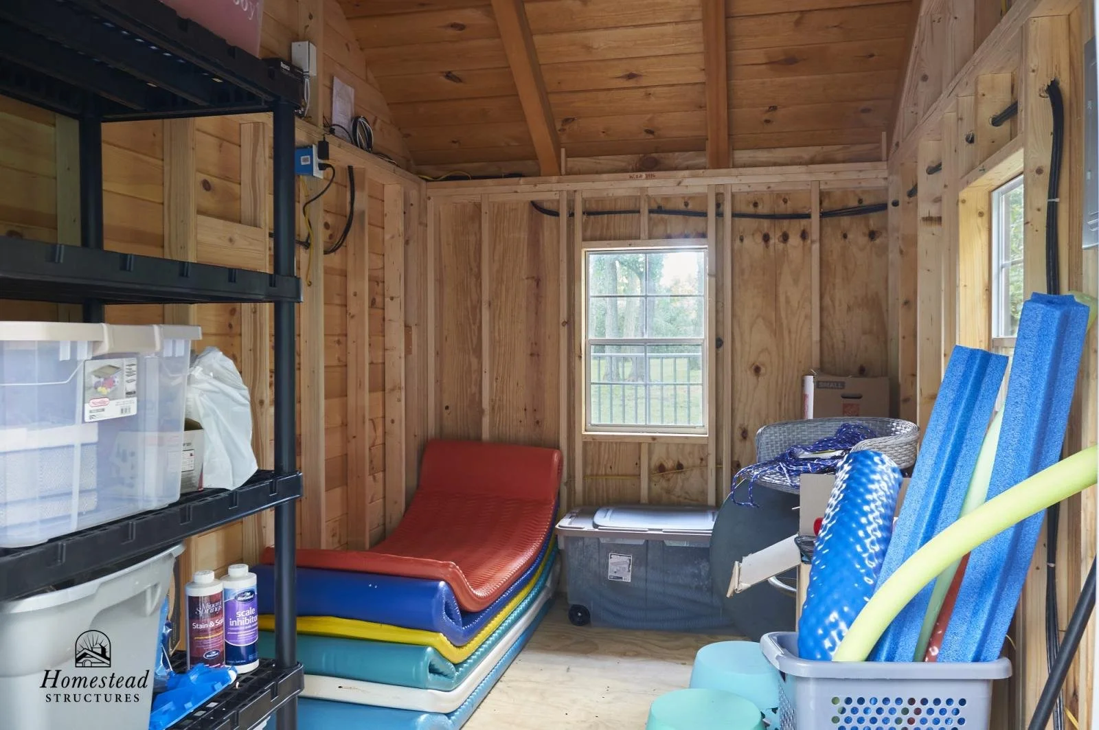 A small shed or storage room with unfinished wooden walls and ceiling, containing colorful pool floats, a plastic storage bin, shelving with various items, two plastic stools, and a window with a view of trees outside.