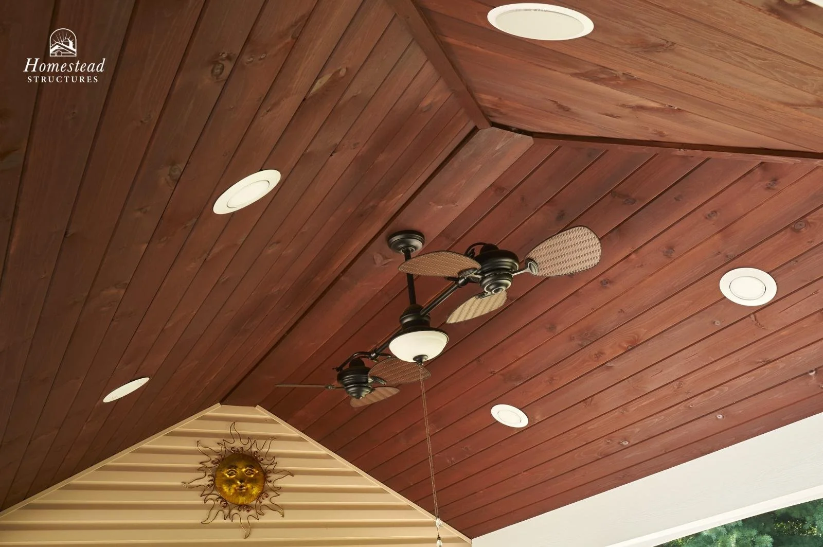 Wood-paneled ceiling with four recessed lights, fan with wicker blades and a frosted glass light, and a sun-shaped wall decoration.