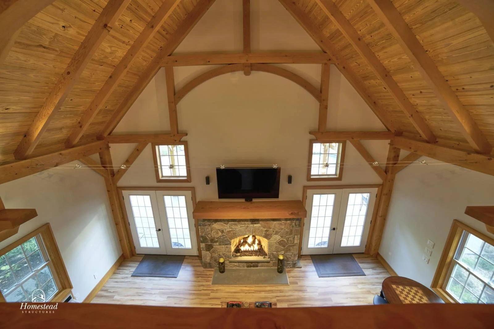 View of a living room with a high wooden ceiling, a stone fireplace with a wood mantel, a mounted TV above the fireplace, and large glass doors leading outside. Two windows flank the doors, and there are two small rugs on the hardwood floor.