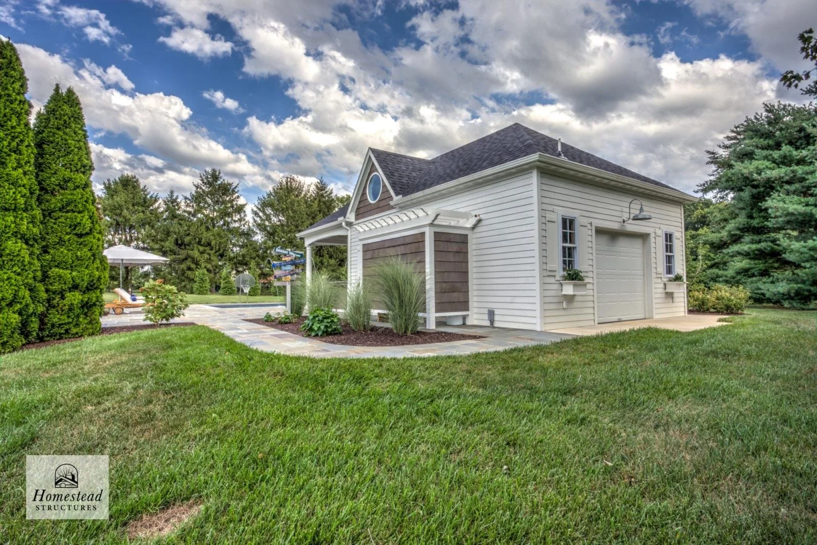 A white garage with a closed door, small windows with flower boxes, a gray roof, and exterior lighting, surrounded by a well-maintained lawn and garden with shrubs and trees, under a partly cloudy sky.