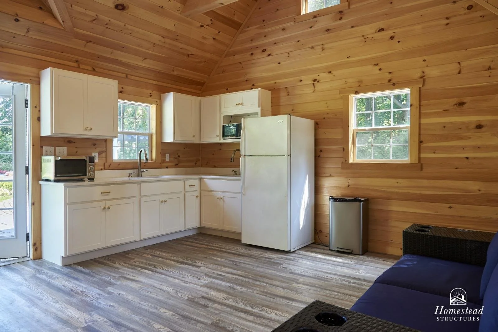 Kitchen with white cabinets, wooden walls, and laminate floors, featuring a microwave, refrigerator, small sink, window, and a dark sofa in the corner.
