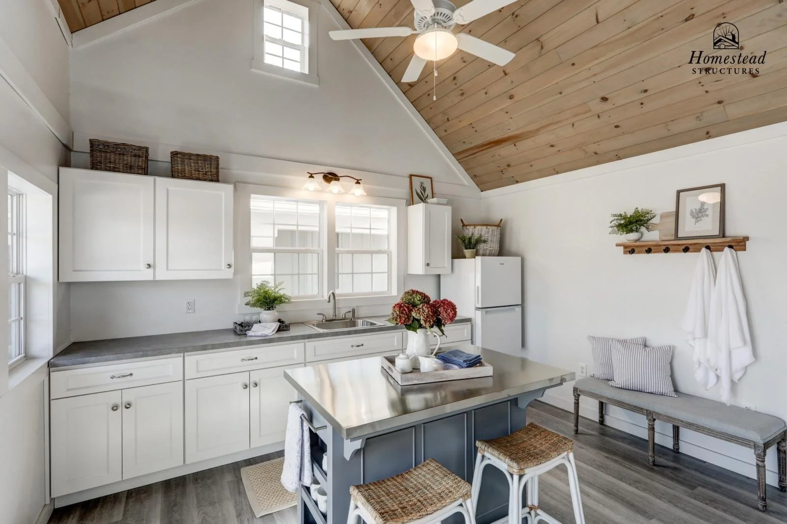 Bright, white kitchen with a vaulted ceiling, wooden accents, and modern decor, counter with flowers and kitchen utensils, window near sink, and a small seating area on the side.