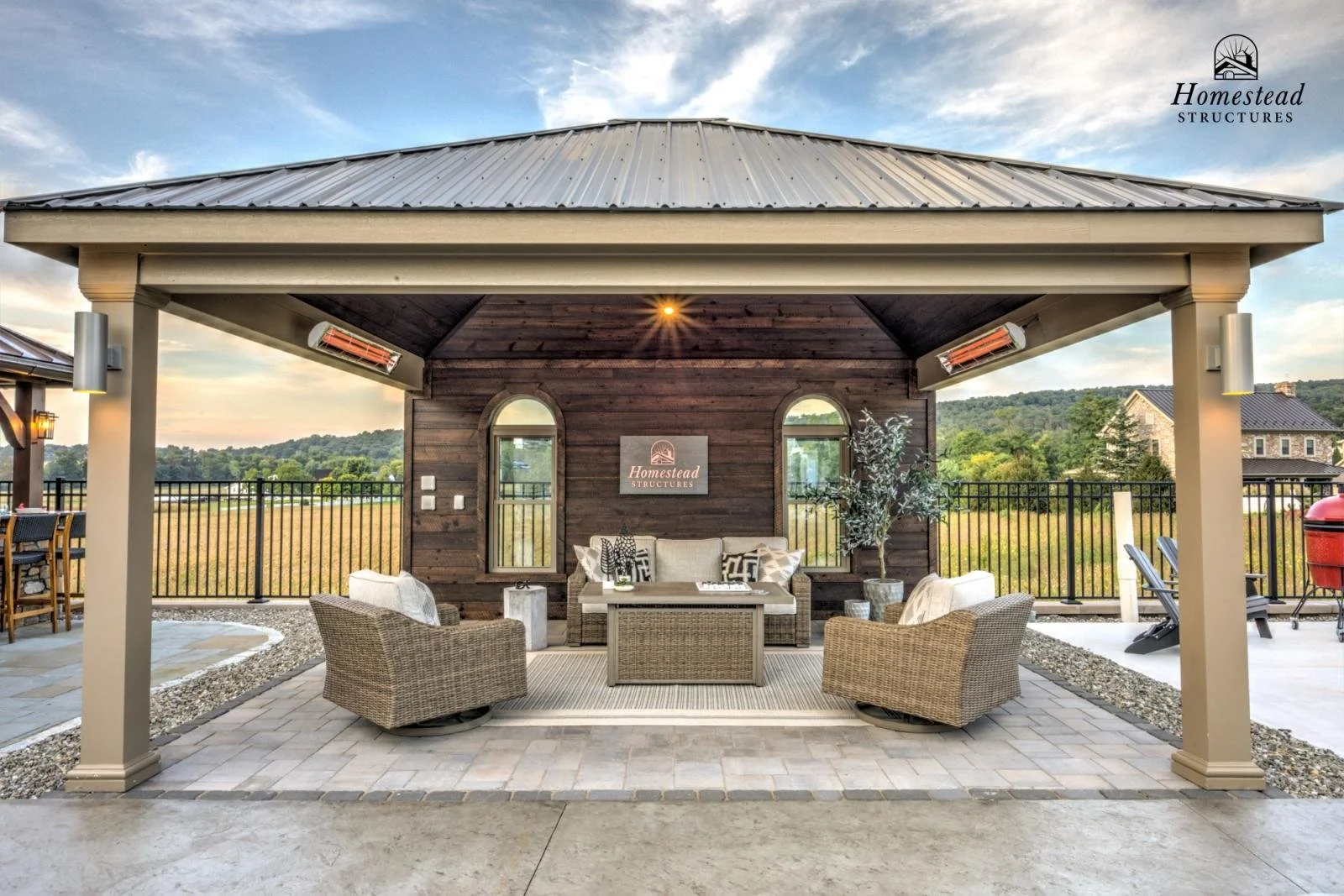 Outdoor covered seating area with wicker chairs, a wood-paneled wall with a sign reading "Homestead Structures," and scenic hills in the background.