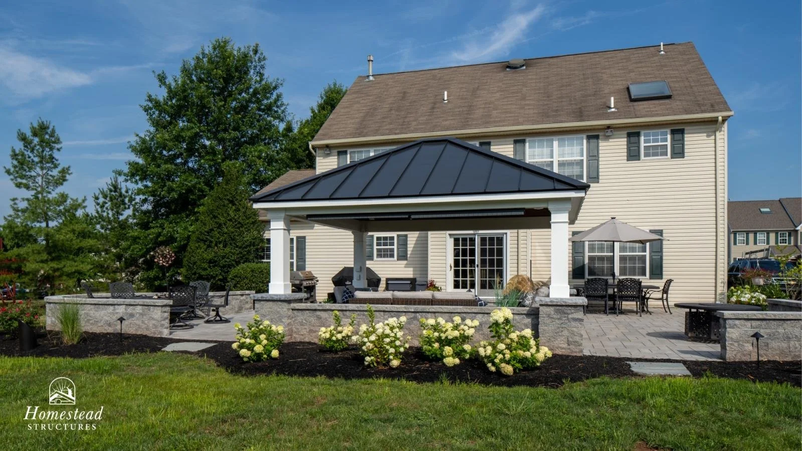 Backyard patio with seating, umbrellas, fireplace, and outdoor kitchen, adjacent to a beige house with green shutters, surrounded by trees and plants under a blue sky.