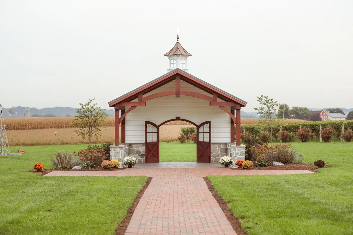 Country-style building with a white wooden exterior, a red gable roof, and a small tower on top, situated in a green lawn with flower beds and a brick pathway leading to open double doors.