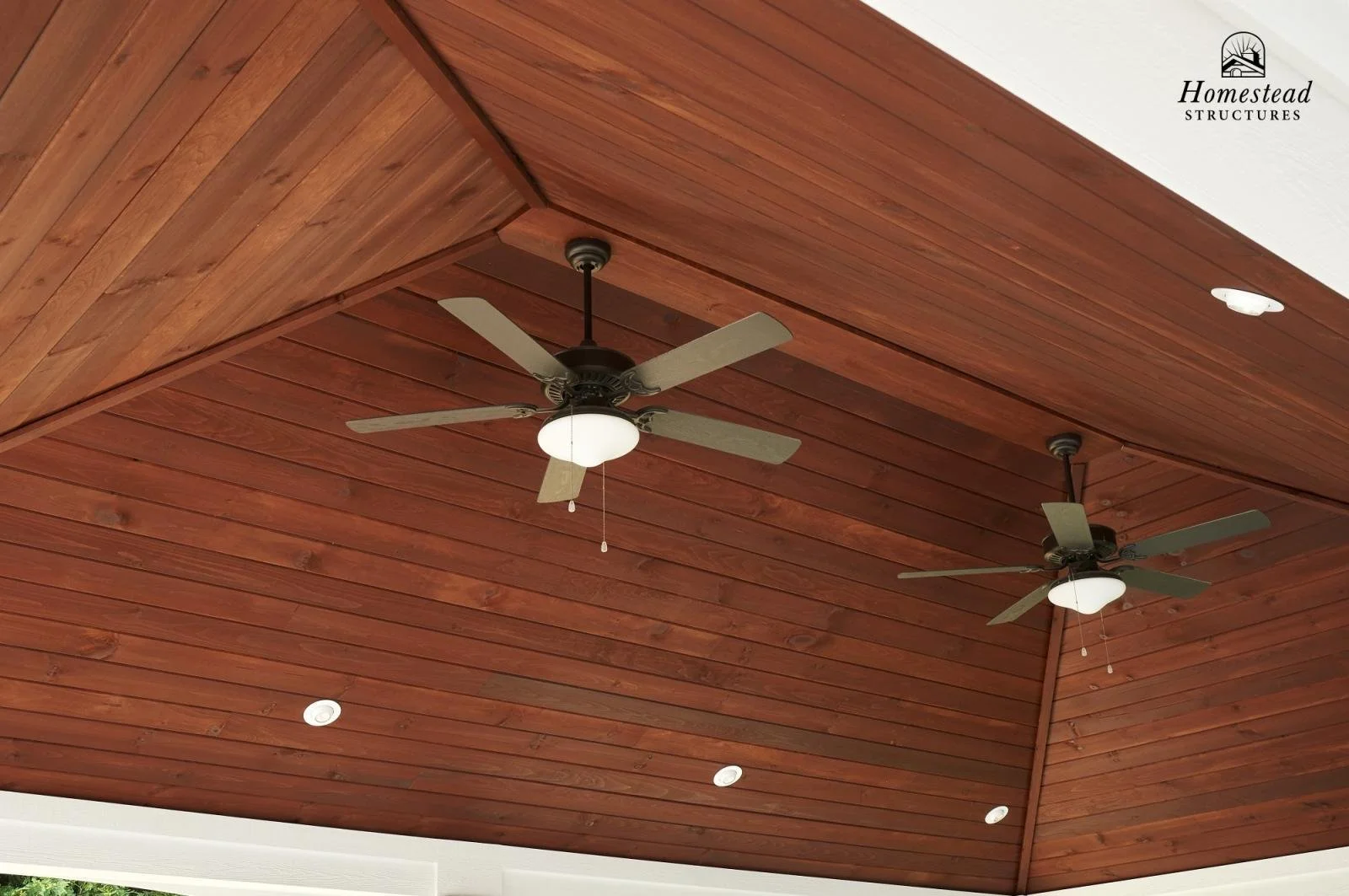 Ceiling of a wooden porch with two ceiling fans and recessed lighting, with the logo 'Homestead Structures' in the top right corner.