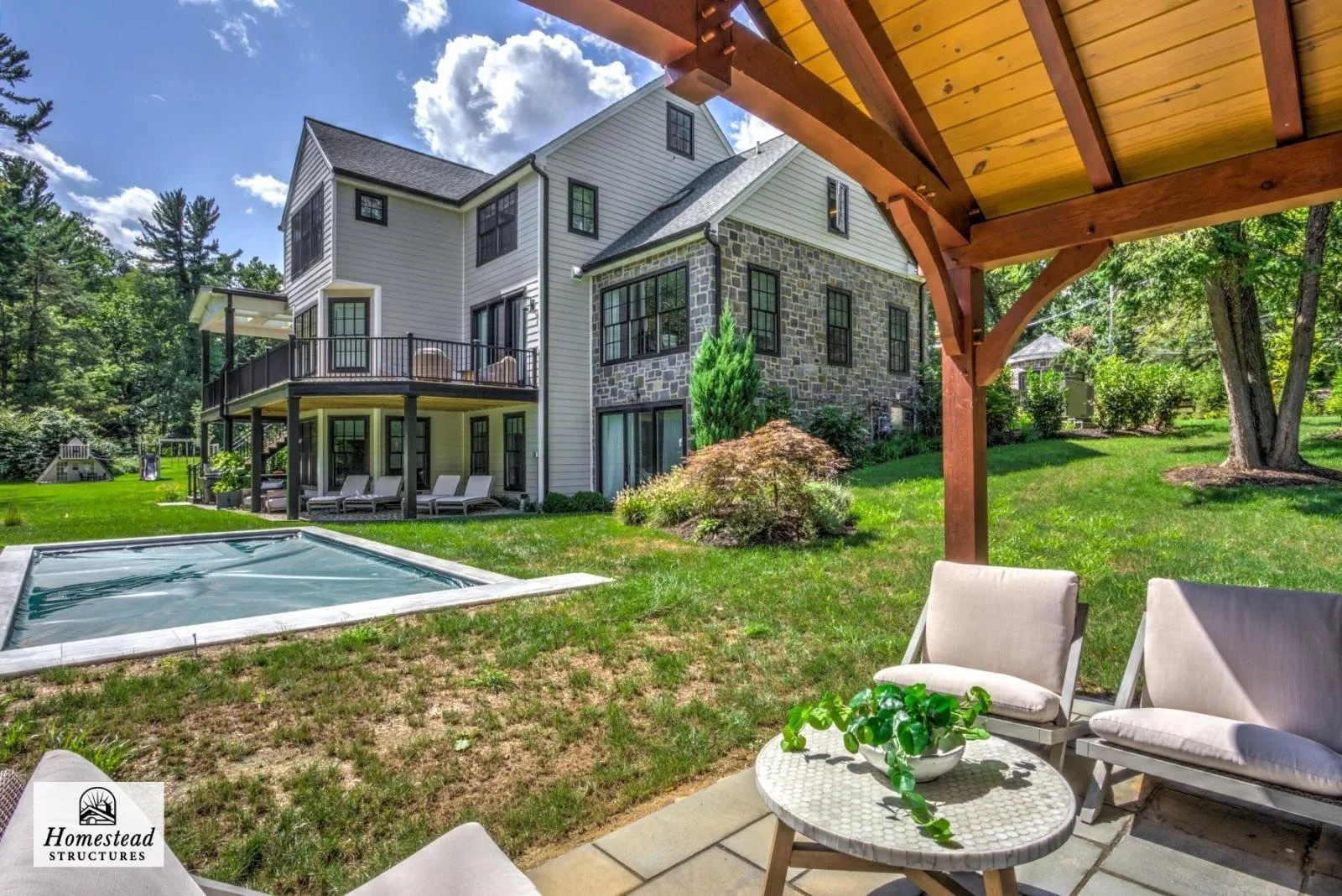 Backyard of a modern house with a deck, swimming pool, patio furniture under a wooden pergola, and lush green landscaping.