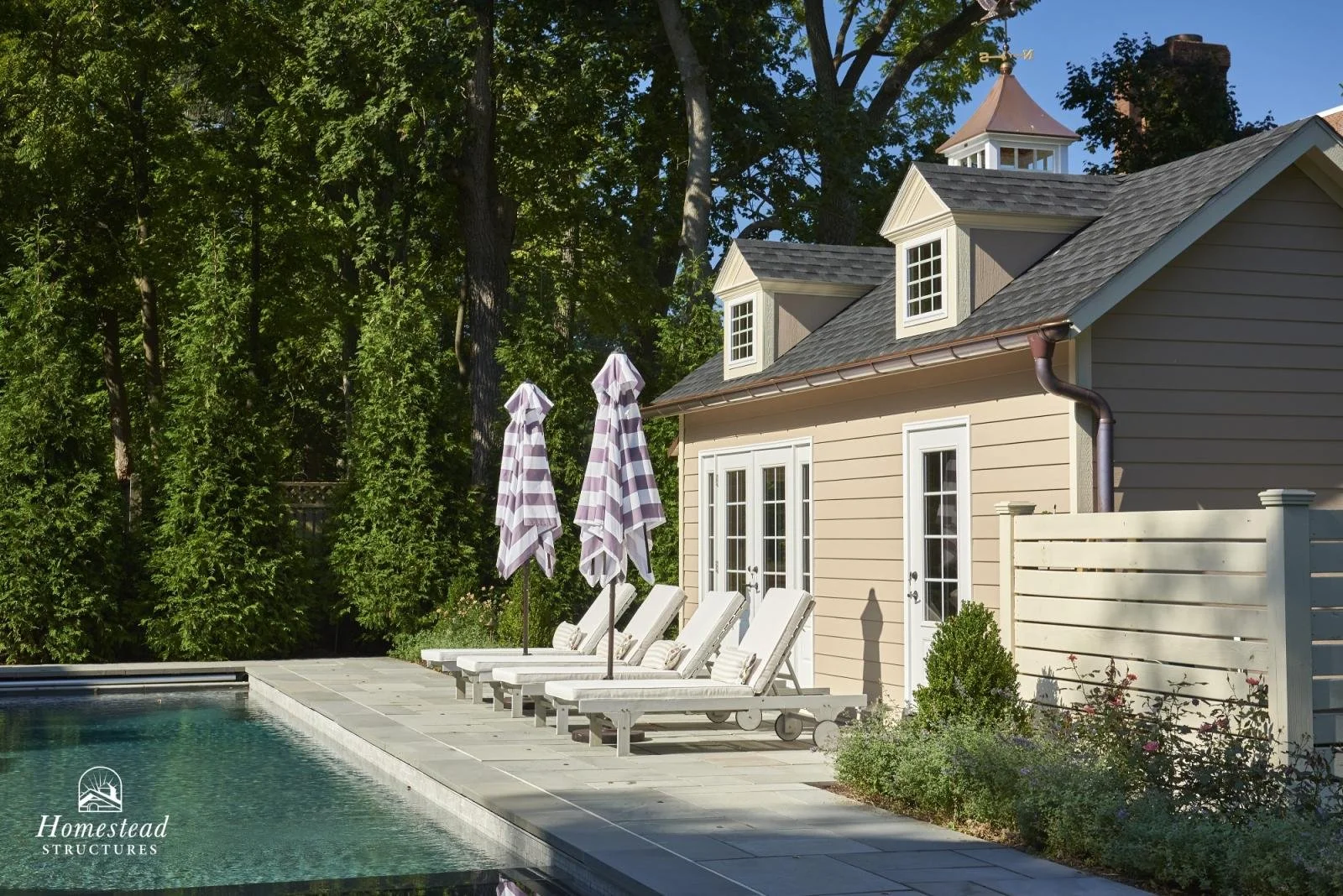 Backyard with swimming pool, white lounge chairs with cushions, and striped umbrellas, beige house with dormer windows, and green trees in the background.