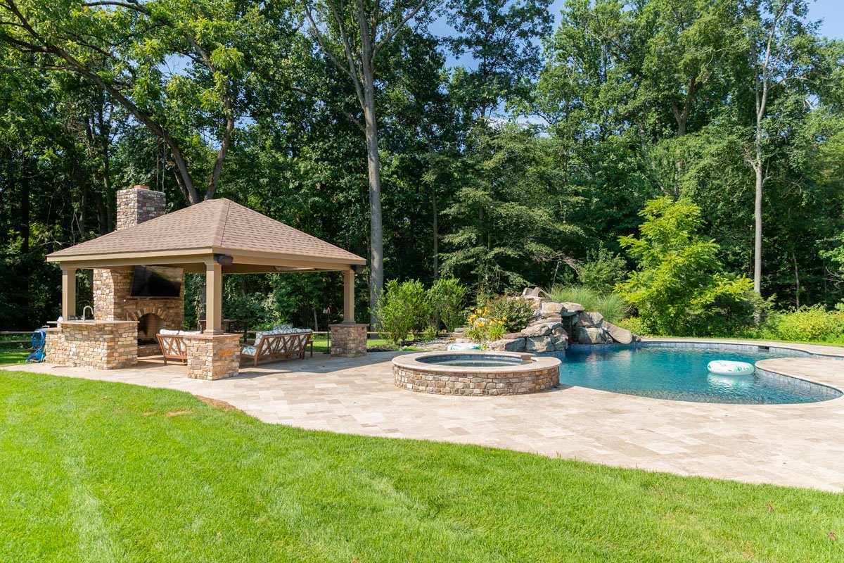 Backyard with a swimming pool, Jacuzzi, and a covered outdoor kitchen with a TV, surrounded by lush green trees.