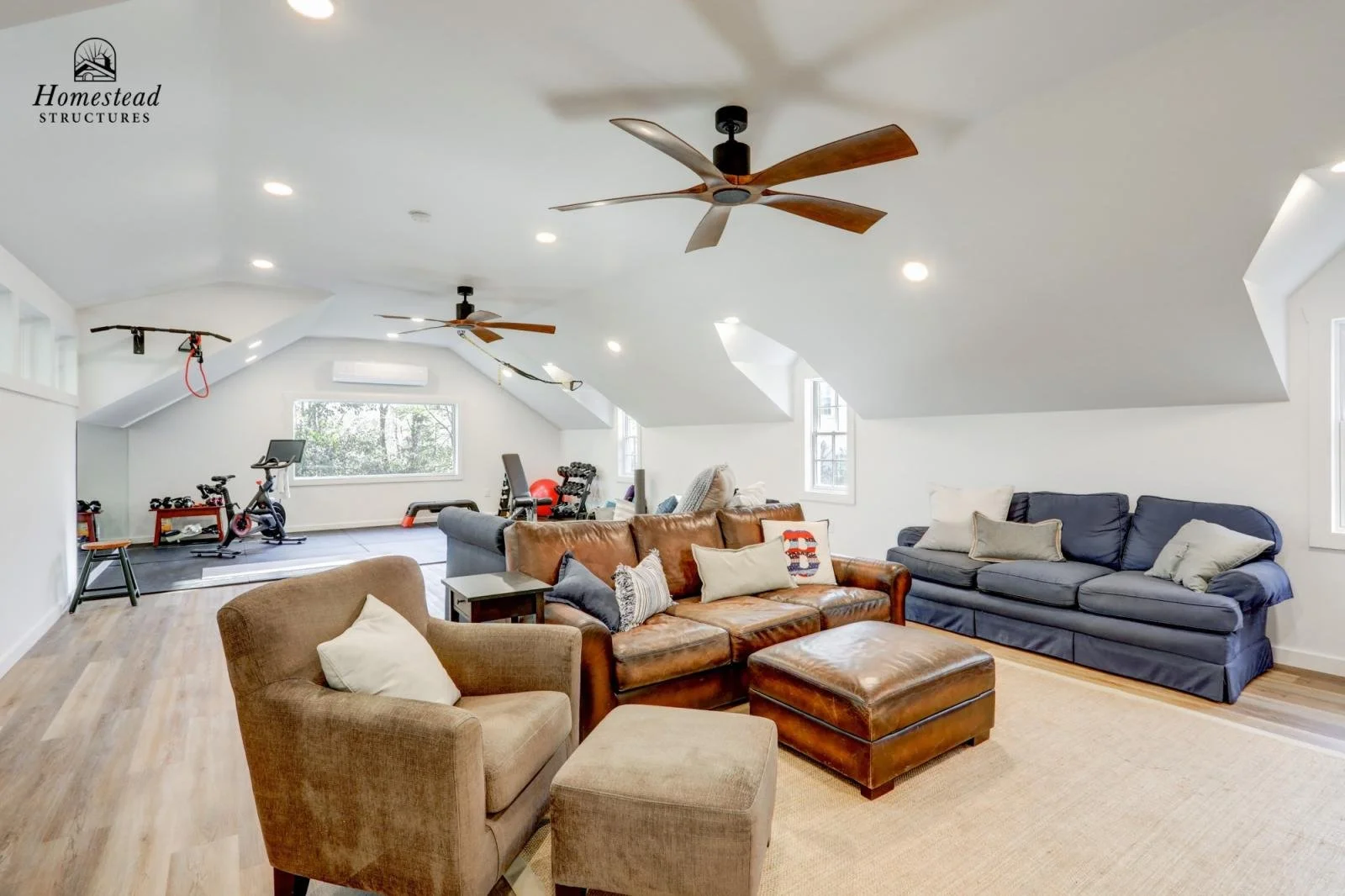 Living room with white walls and ceiling, ceiling fans, windows, brown leather sofa, blue sofa, beige armchair with ottoman, and exercise equipment in the background.
