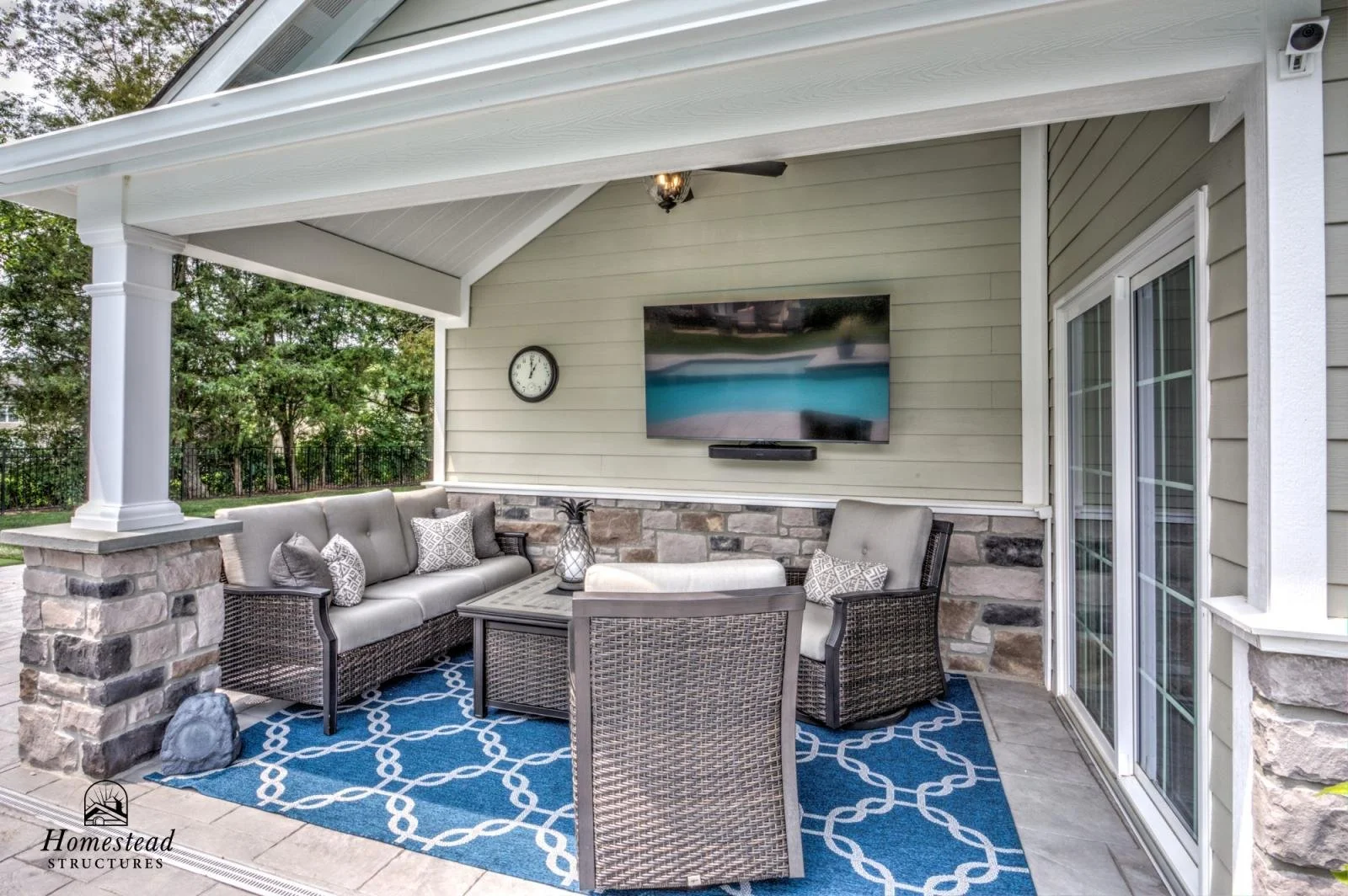 Outdoor patio with rattan furniture, cushions, and a blue geometric rug under a screened porch with beige siding, stone accents, a wall-mounted TV, and a sliding glass door.